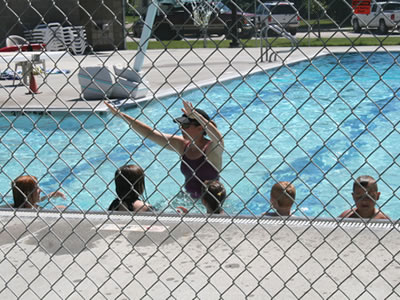 There is a chain link fabric fence that installed around the swimming pool, with a lady and five boys in the pool.